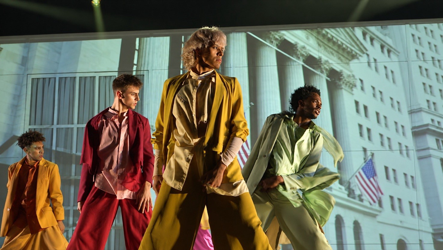 AMDA dancers in red, yellow and pale-green costumes mid-pose in The Glory Hour, before a projected classical building facade