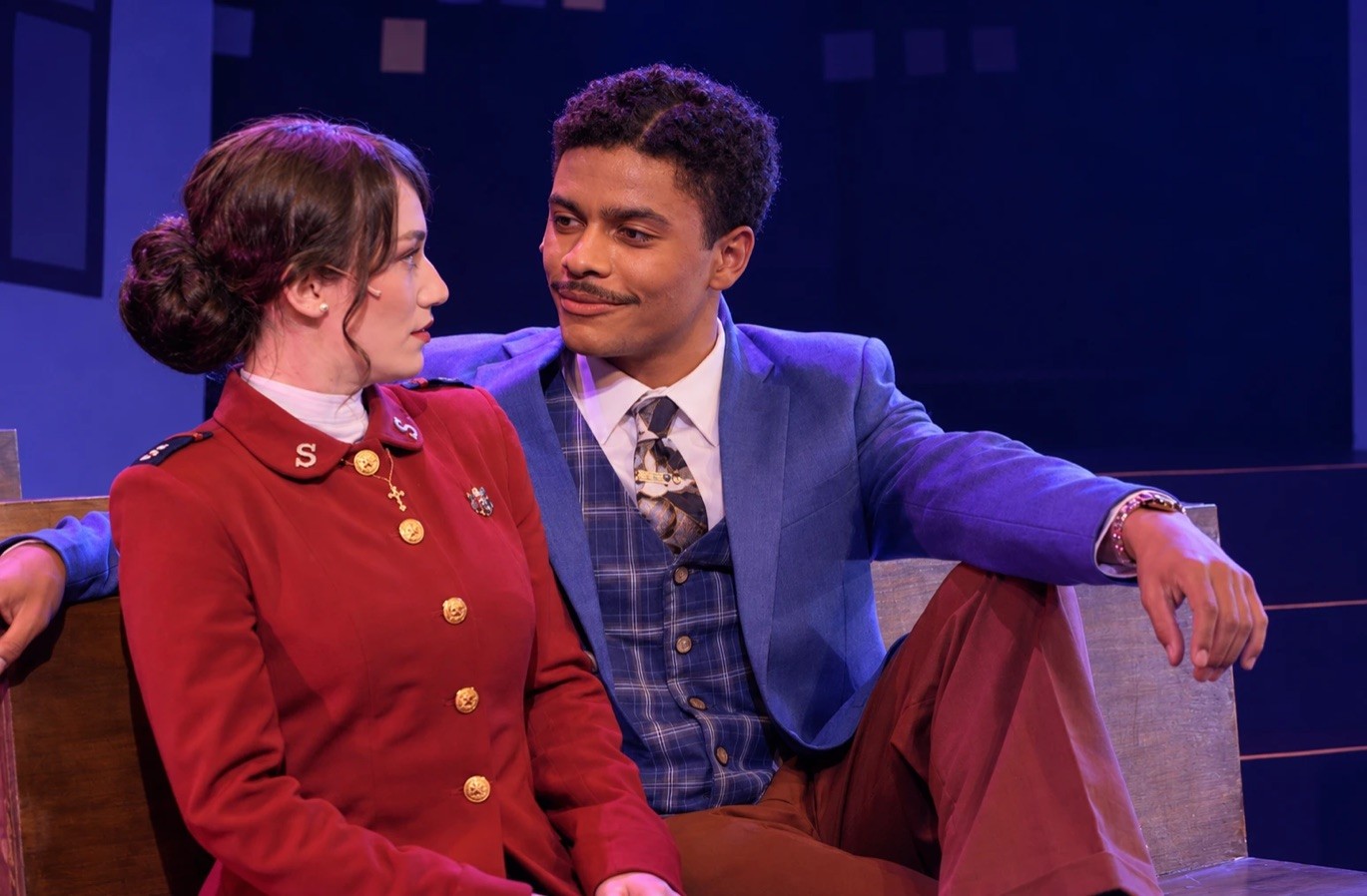 Two AMDA students on stage in Guys and Dolls, he in a blue jacket leaning back on a bench and smiling at her in a red Salvation Army uniform