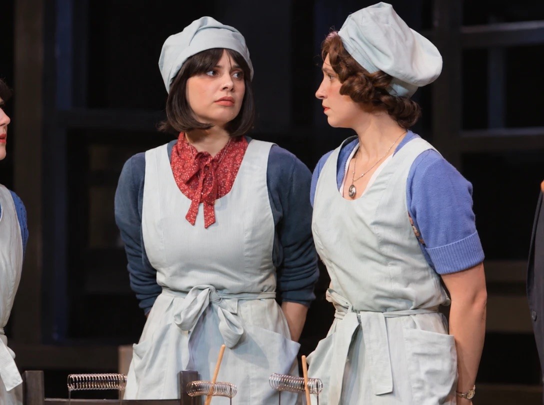 Two AMDA actors in pale-blue factory aprons and caps in Radium Girls, looking at each other